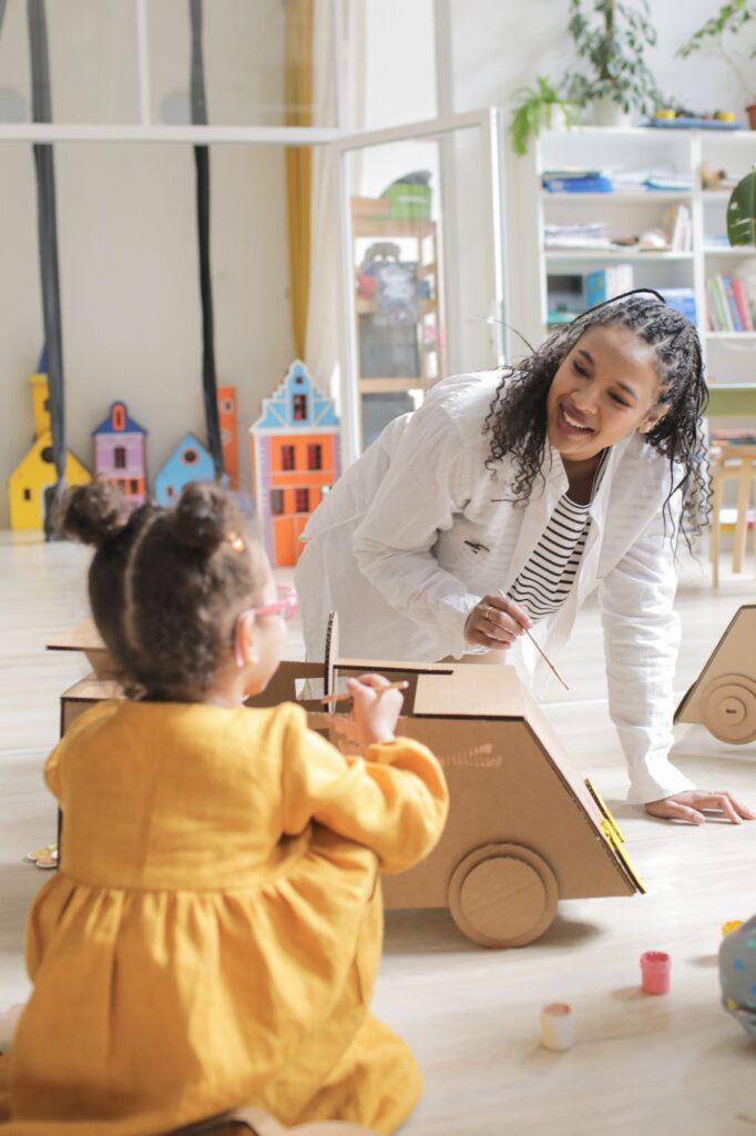 A smiling therapist painting a cardboard car with a toddler